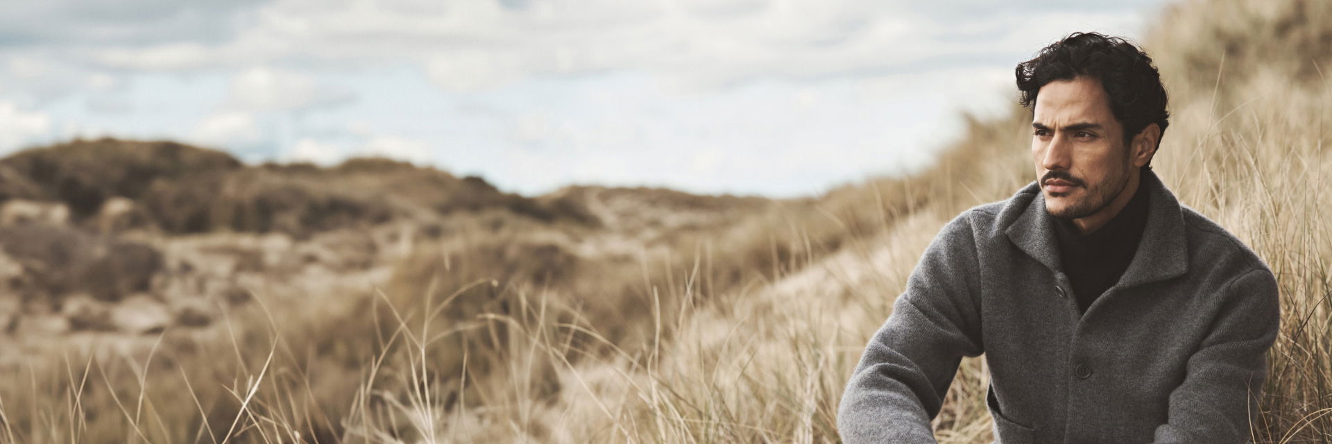 Sitzender Mann mit schwarzem Rollkragenpullover, grauer Kaschmirjacke und schwarzer Hose in Dünenlandschaft, nachdenklich blickend. Sitzender Mann mit schwarzem Rollkragenpullover, grauer Kaschmirjacke und schwarzer Hose in Dünenlandschaft, nachdenklich blickend.