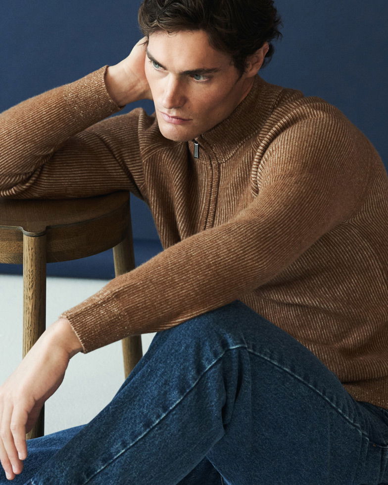 Mann mit braunem Strickpullover und dunkler Jeans sitzt nachdenklich auf dem Boden, Arm abgestützt auf hölzernem Stuhl.