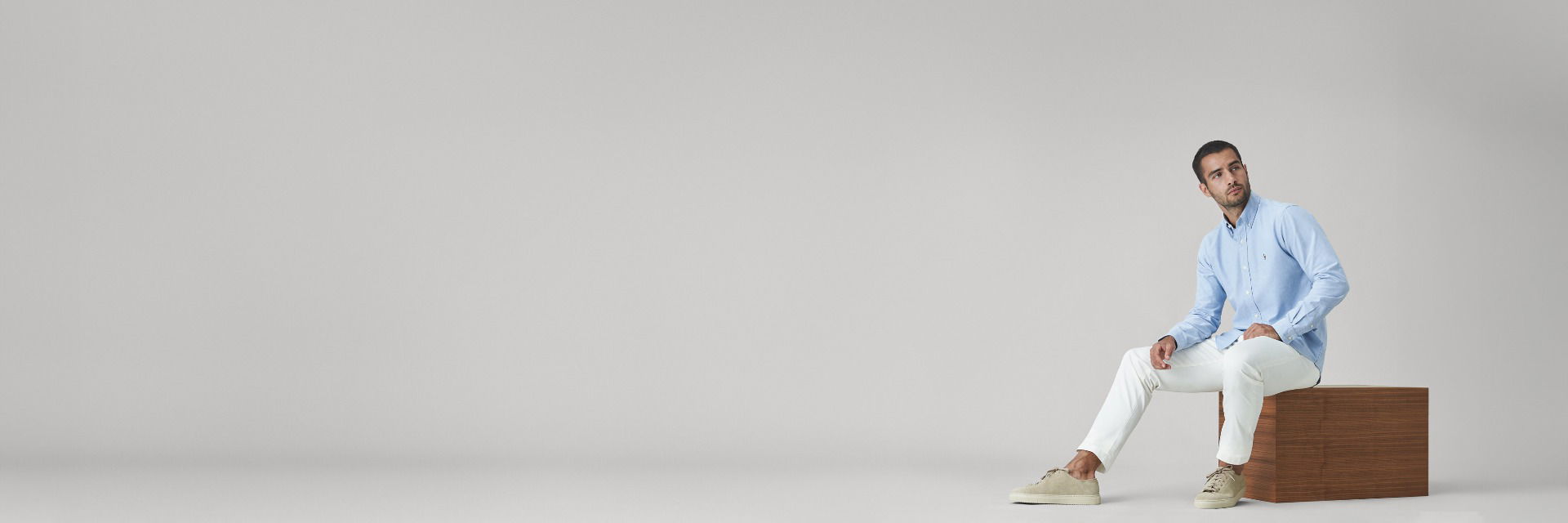 Mann sitzt auf hölzernem Würfel, trägt hellblaues Hemd, weiße Chinohose und beige Sneaker, schaut nach rechts. Mann sitzt auf hölzernem Würfel, trägt hellblaues Hemd, weiße Chinohose und beige Sneaker, schaut nach rechts.