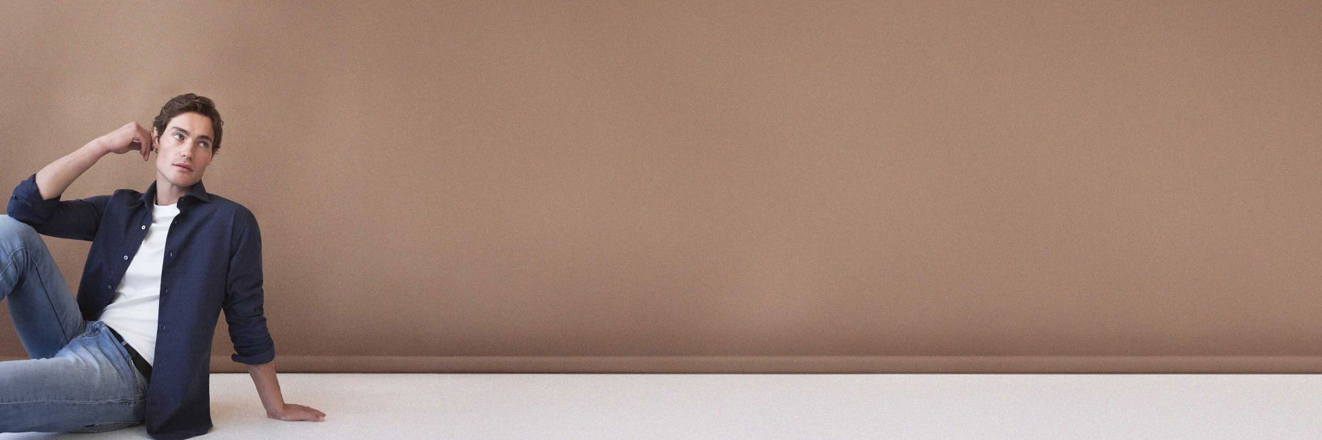 Sitzender Mann mit blauem Flanellhemd, weißem T-Shirt, blauen Jeans und schwarzem Gürtel vor braunem Hintergrund. Sitzender Mann mit blauem Flanellhemd, weißem T-Shirt, blauen Jeans und schwarzem Gürtel vor braunem Hintergrund.