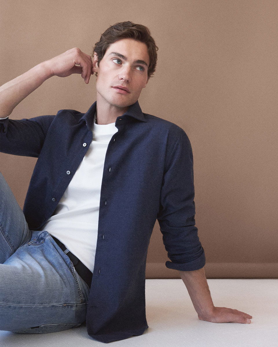 Junger Mann sitzt vor beige Wand, trägt blaues Flanellhemd, weißes T-Shirt, hellblaue Jeans und schwarzen Gürtel.