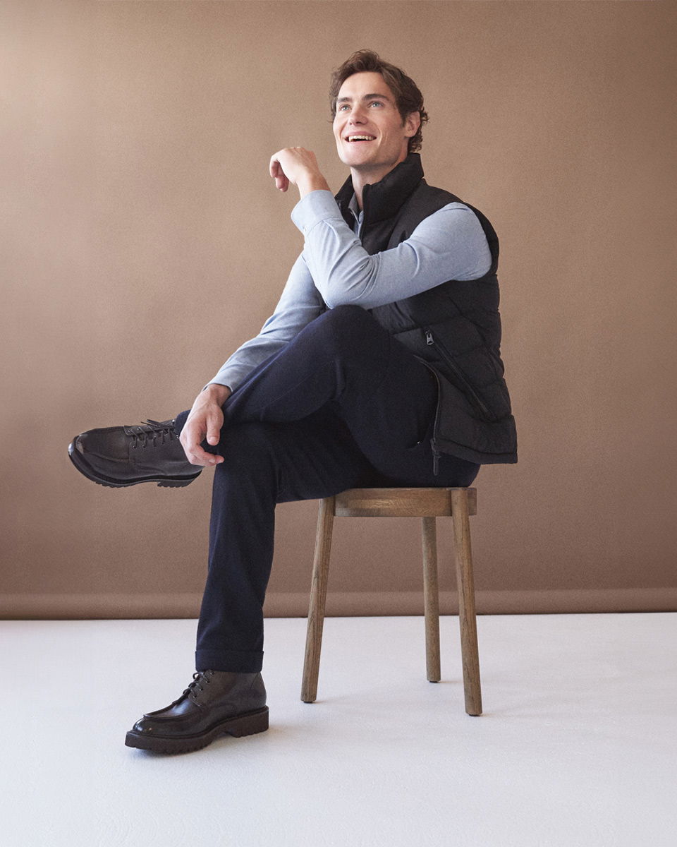 Sitzender Mann mit blauem Hemd, schwarzer Weste, blauer Hose und braunen Schnürboots auf Holzhocker vor braunem Hintergrund.