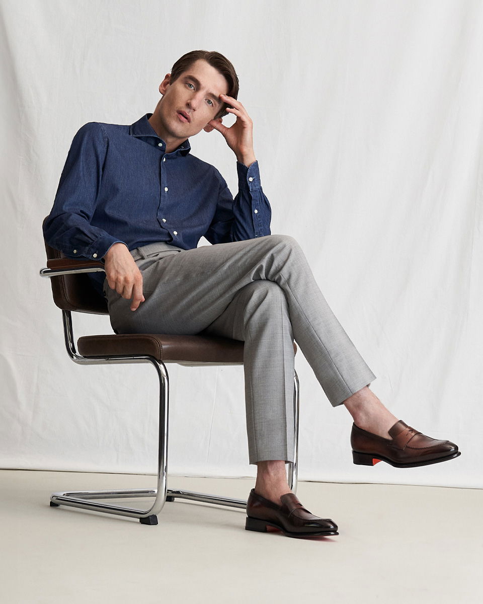 Mann sitzt auf braunem Stuhl, trägt dunkelblaues Hemd, graue Hose und braune Loafer, linker Arm an Kopf gestützt. Mann sitzt auf braunem Stuhl, trägt dunkelblaues Hemd, graue Hose und braune Loafer, linker Arm an Kopf gestützt.