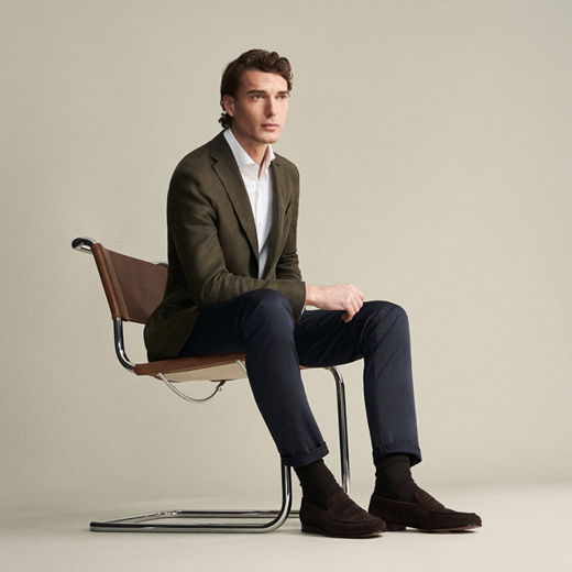 Mann sitzt auf Stuhl, trägt olivgrünes Sakko, weißes Hemd, navy Hose mit Krempel, braune Loafer und dunkle Socken. Mann sitzt auf Stuhl, trägt olivgrünes Sakko, weißes Hemd, navy Hose mit Krempel, braune Loafer und dunkle Socken.