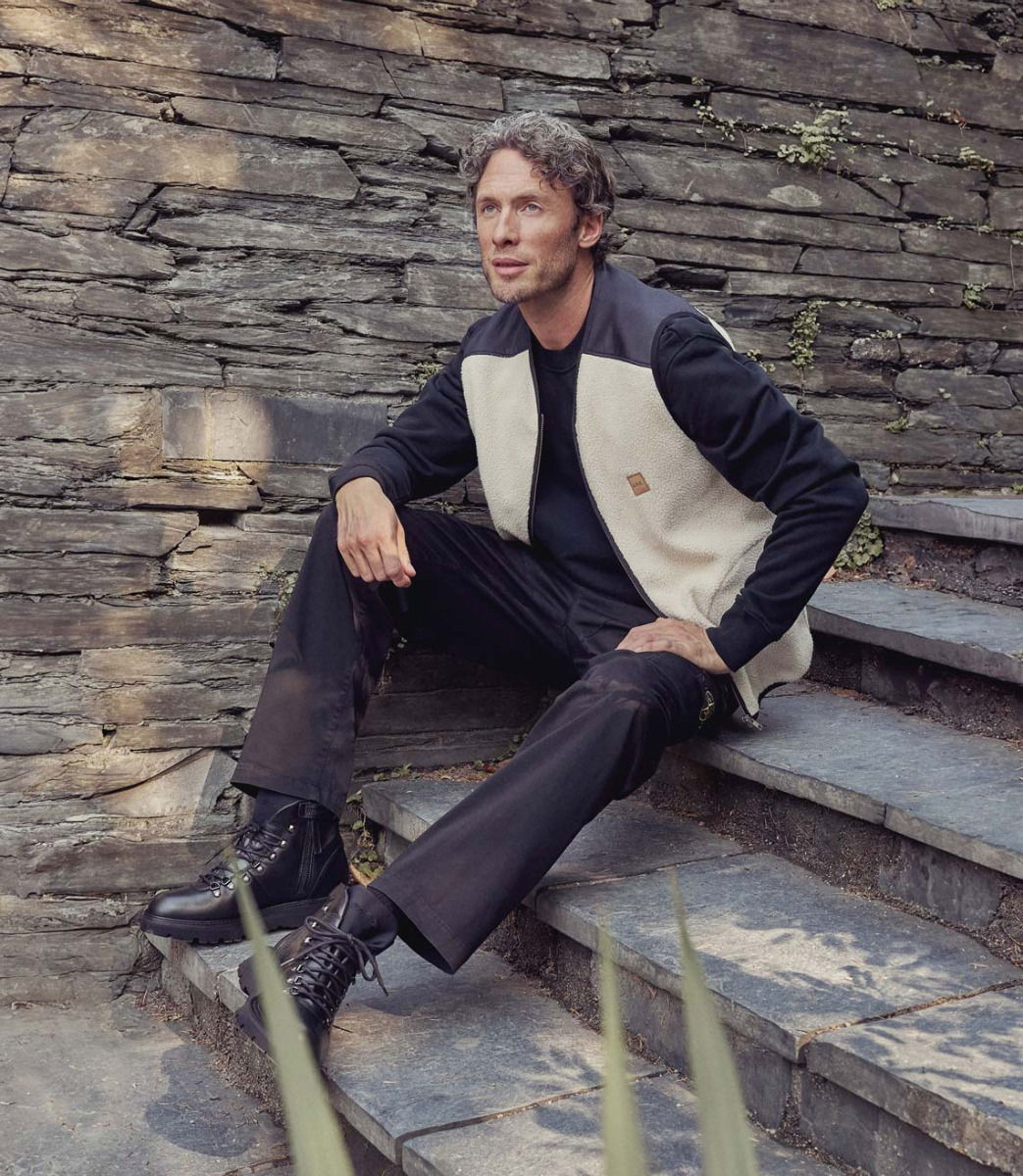 Mann mit lockigem Haar sitzt auf steiniger Treppe, trägt schwarze Hose, schwarzen Pullover, beige Weste und schwarze Boots.