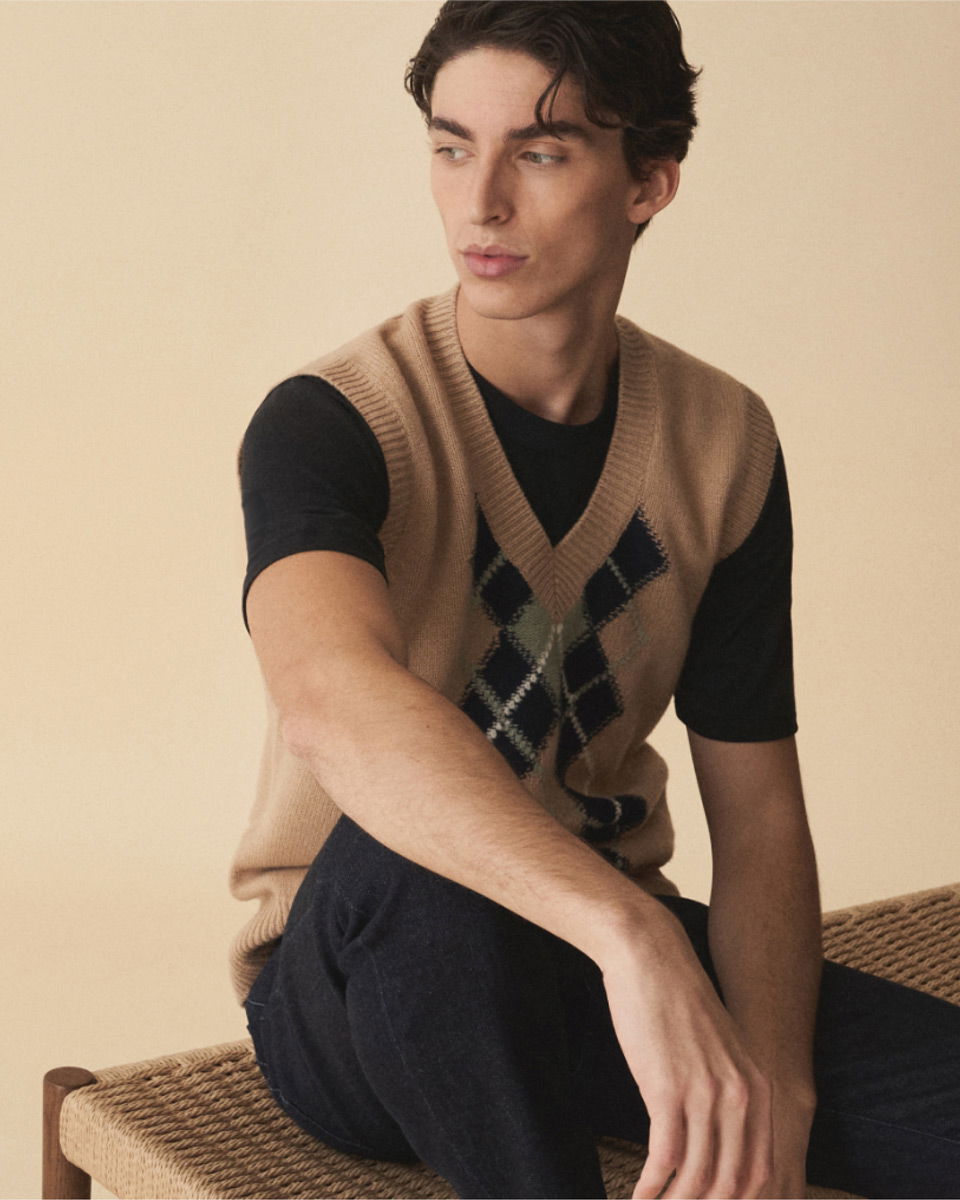 Junger Mann sitzt auf einem geflochtenen Stuhl, trägt schwarzen T-Shirt und braunen Pullunder mit Rautenmuster.