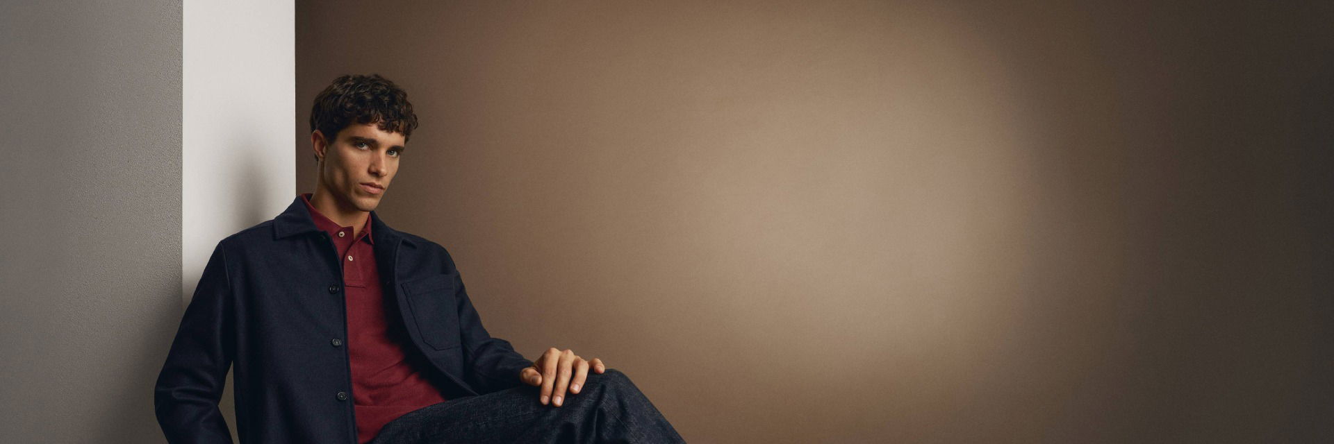 Sitzender Mann mit dunklem Overshirt, dunkelrotem Poloshirt und dunkelblauer Jeans lehnt an weißer Wand vor beigem Hintergrund. Sitzender Mann mit dunklem Overshirt, dunkelrotem Poloshirt und dunkelblauer Jeans lehnt an weißer Wand vor beigem Hintergrund.