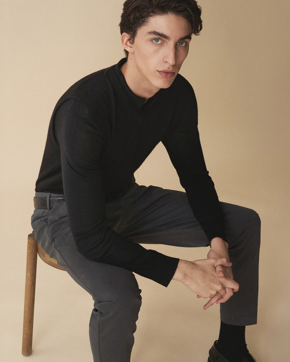 Junger Mann sitzt auf Holzstuhl, trägt schwarzen Pullover, schwarzen Pullunder, dunkelgraue Hose und schwarze Schuhe.