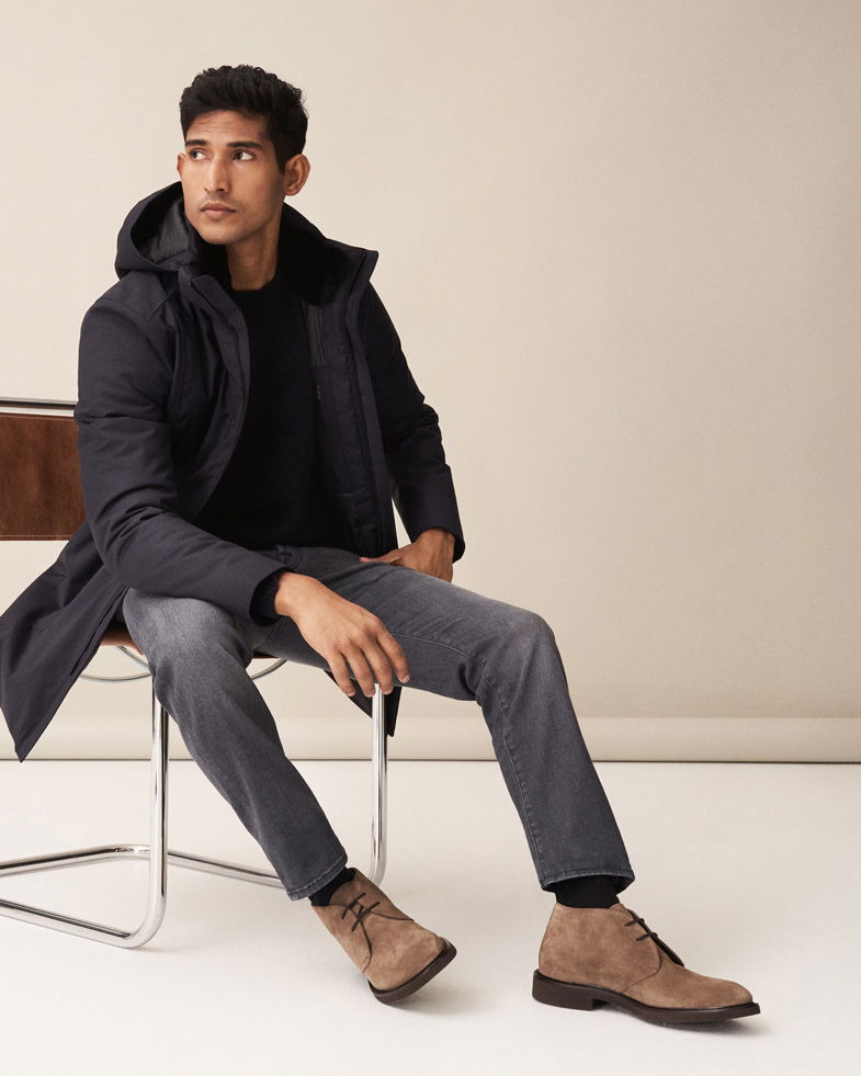 Sitzender Mann mit schwarzen Daunenmantel, grauer Jeans, dunkelblauem Pullover und beigen Desert-Boots vor hellem Hintergrund.