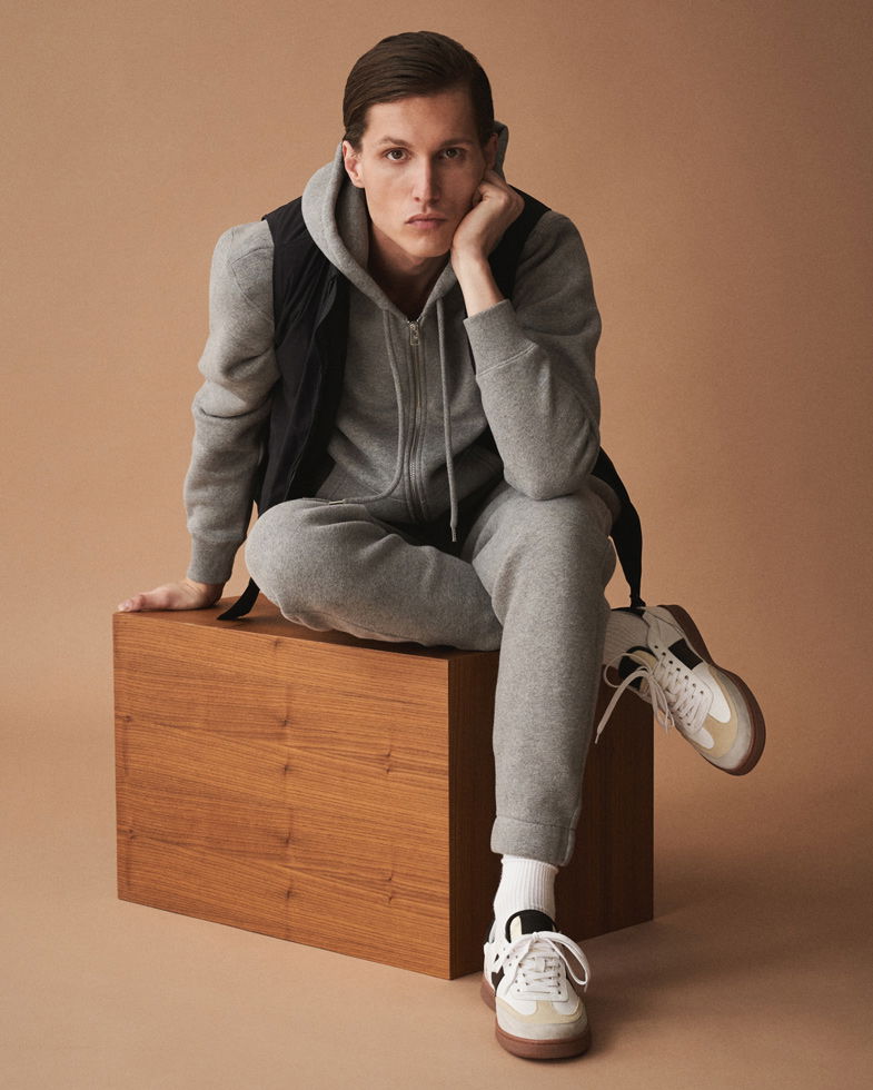 Junger Mann sitzt auf Holzkiste, trägt grauen Jogginganzug, schwarze Weste und weiße Retro-Sneaker, vor beige Hintergrund.