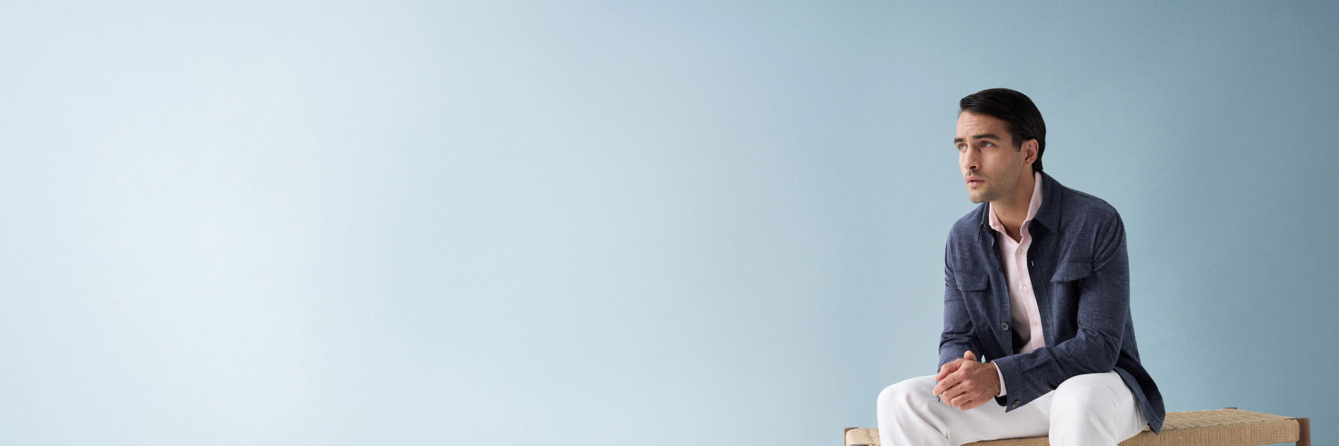Sitzender Mann mit dunkelblauem Overshirt, rosa Hemd und weißer Hose vor hellblauem Hintergrund, nach rechts schauend. Sitzender Mann mit dunkelblauem Overshirt, rosa Hemd und weißer Hose vor hellblauem Hintergrund, nach rechts schauend.
