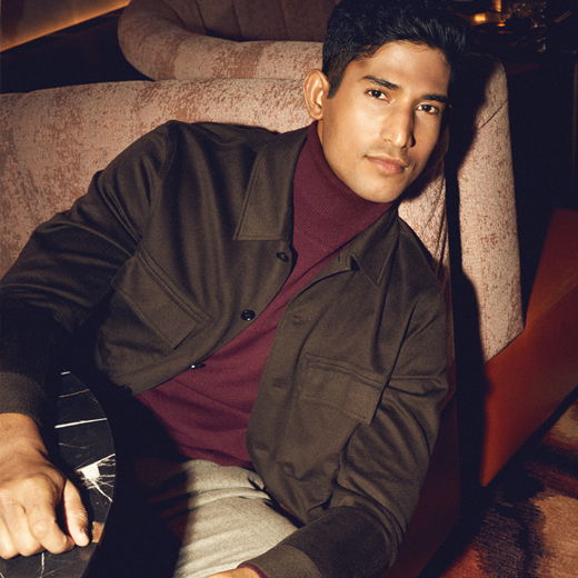 Junger Mann sitzt entspannt auf rosafarbenem Sofa, trägt braune Overshirt-Jacke, bordeauxfarbenen Rollkragenpullover und beige Hose. Junger Mann sitzt entspannt auf rosafarbenem Sofa, trägt braune Overshirt-Jacke, bordeauxfarbenen Rollkragenpullover und beige Hose.