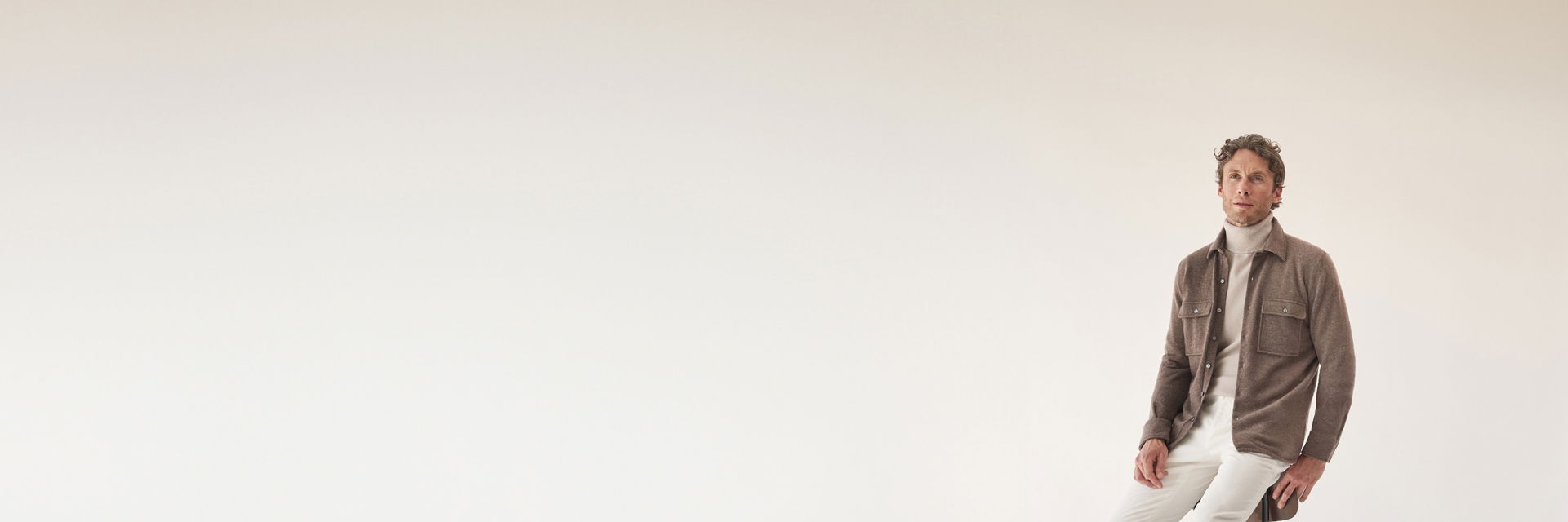 Mann sitzt vor hellen Hintergrund, trägt beigen Rollkragenpullover, braunes Overshirt und cremefarbene Hose, schaut frontal. Mann sitzt vor hellen Hintergrund, trägt beigen Rollkragenpullover, braunes Overshirt und cremefarbene Hose, schaut frontal.