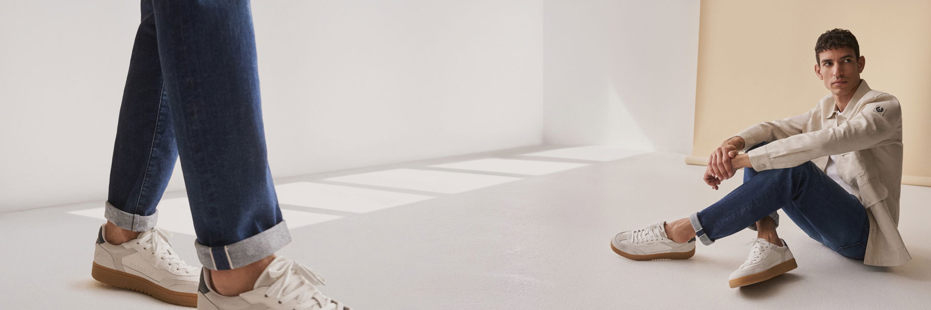 Sitzender Mann mit beige Overshirt, dunkelblauer Selvedge Denim Jeans und weißen Sneakern vor hellbeigem Hintergrund. Sitzender Mann mit beige Overshirt, dunkelblauer Selvedge Denim Jeans und weißen Sneakern vor hellbeigem Hintergrund.