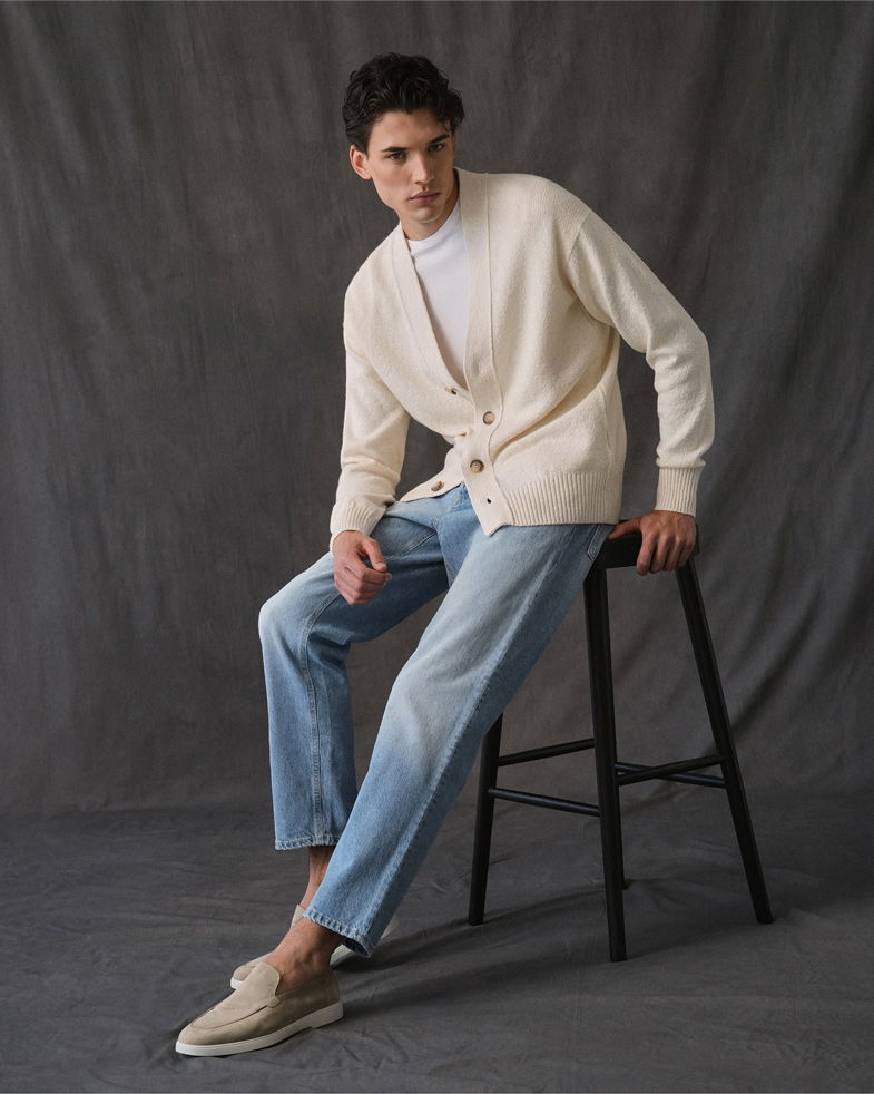 Junger Mann sitzt auf schwarzem Hocker, trägt weißen Cardigan, weißes Shirt, hellblaue Jeans und beige Loafer.