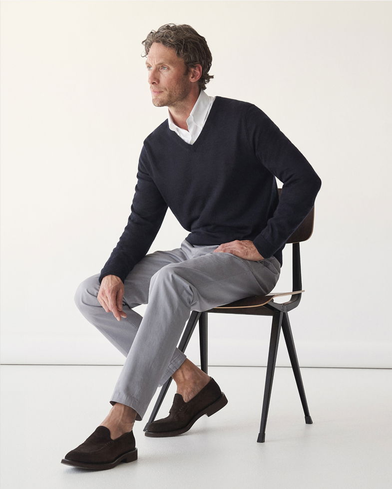 Mann sitzt auf Stuhl, trägt schwarzen V-Neck-Pullover, weißes Hemd, hellgraue Hose und braune Loafer, schaut seitlich. Mann sitzt auf Stuhl, trägt schwarzen V-Neck-Pullover, weißes Hemd, hellgraue Hose und braune Loafer, schaut seitlich.