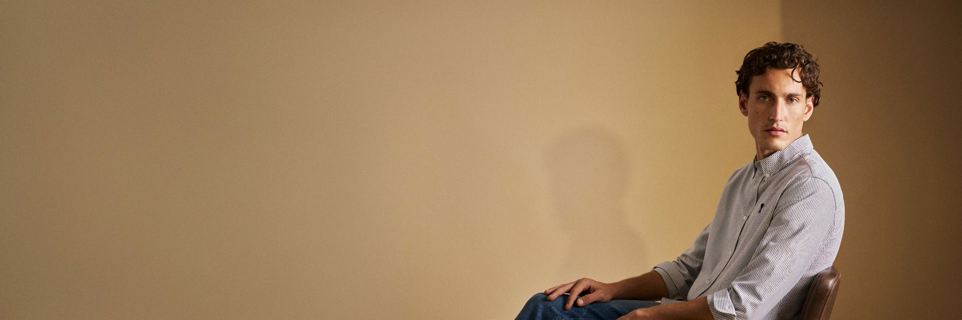 Sitzender Mann mit blau-weiß gestreiftem Hemd, hochgekrempelten Ärmeln und blauer Jeans vor beigem Hintergrund. Sitzender Mann mit blau-weiß gestreiftem Hemd, hochgekrempelten Ärmeln und blauer Jeans vor beigem Hintergrund.