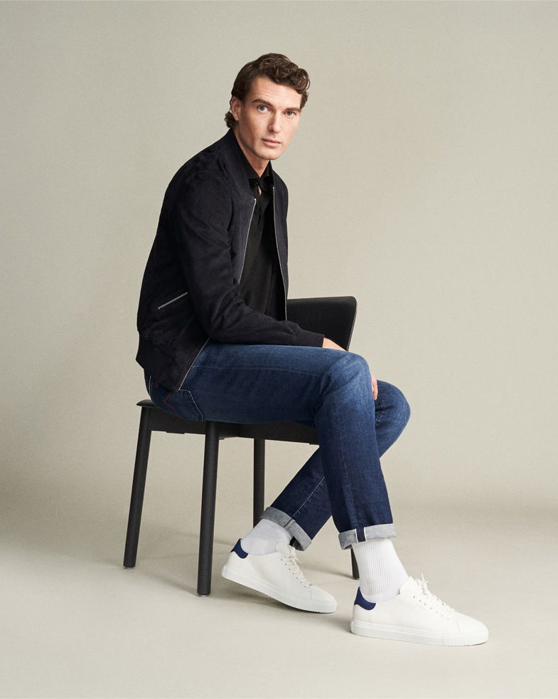 Sitzender Mann mit schwarzer Jacke, schwarzem Shirt, gekrempelter blauer Jeans, weißen Sneakern und weißen Socken vor neutralem Hintergrund. Sitzender Mann mit schwarzer Jacke, schwarzem Shirt, gekrempelter blauer Jeans, weißen Sneakern und weißen Socken vor neutralem Hintergrund.