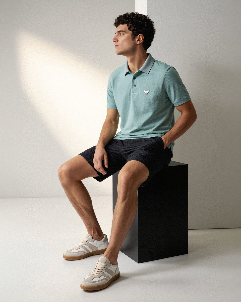 Junger Mann sitzt auf schwarzem Würfel, trägt salbeigrünes Poloshirt, schwarze Shorts und weiße Sneaker mit grauen Details.
