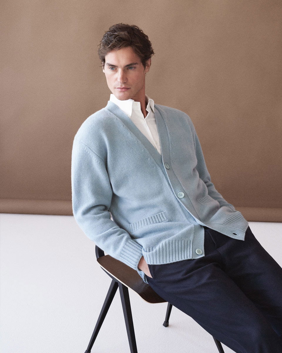 Sitzender Mann mit weißem Hemd, hellblauem Cardigan und dunkelblauer Hose vor schlichtem braunem Hintergrund.