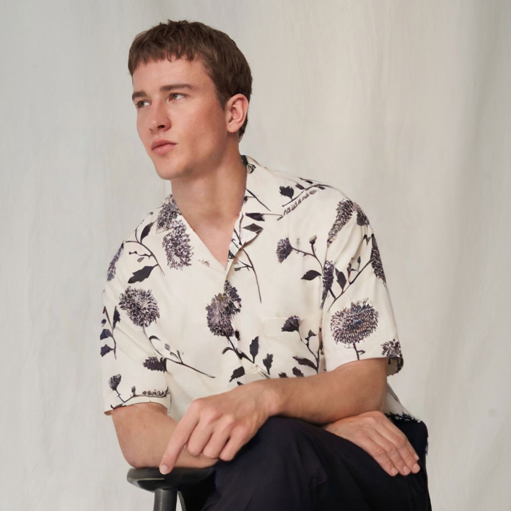 Junger Mann sitzt auf Hocker, trägt weißes Kurzarmhemd mit braunem Blumenmuster und schwarze Hose, blickt nach links. Junger Mann sitzt auf Hocker, trägt weißes Kurzarmhemd mit braunem Blumenmuster und schwarze Hose, blickt nach links.
