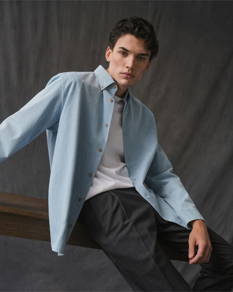 Junger Mann sitzt vor grauer Studiowand, trägt hellblaues Jeanshemd, weißes T-Shirt und schwarze Hose, Blick nach vorne.