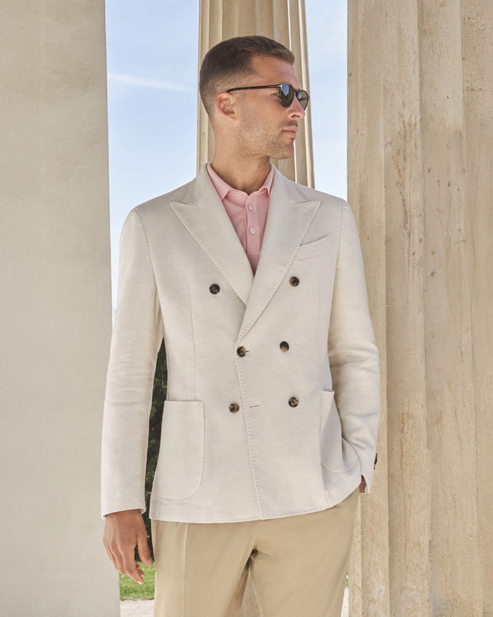 Eleganter Mann mit beige Hose, cremefarbenem Doppelreiher-Sakko und rosa Hemd, trägt Sonnenbrille, vor Säulen stehend.