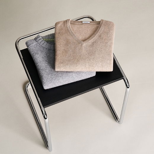 Zwei gefaltete Herrenpullover, beige und grau, liegen auf einem schwarzen Hocker mit verchromtem Gestell. Zwei gefaltete Herrenpullover, beige und grau, liegen auf einem schwarzen Hocker mit verchromtem Gestell.