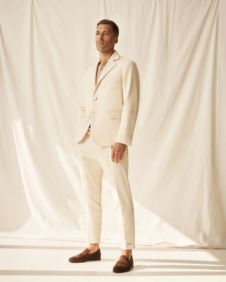 Mann steht vor beigem Hintergrund, trägt cremefarbenen Anzug, hellbraunes Strickpolo und braune Penny-Loafer-Schuhe.