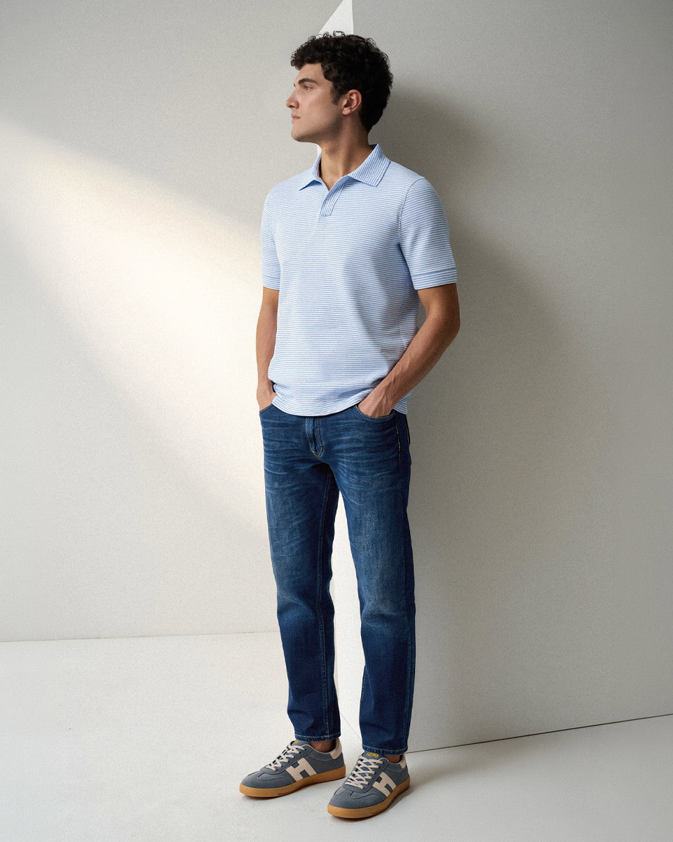 Junger Mann steht seitlich, trägt hellblau gestreiftes Strickpolo, blaue Jeans und graublaue Sneaker mit weißen Details.
