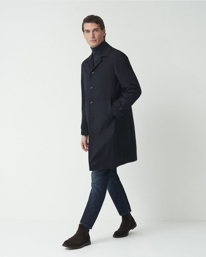 Mann steht vor weißem Hintergrund, trägt schwarzen Kurzmantel, blauen Rollkragenpullover, blaue Jeans und schwarze Boots.