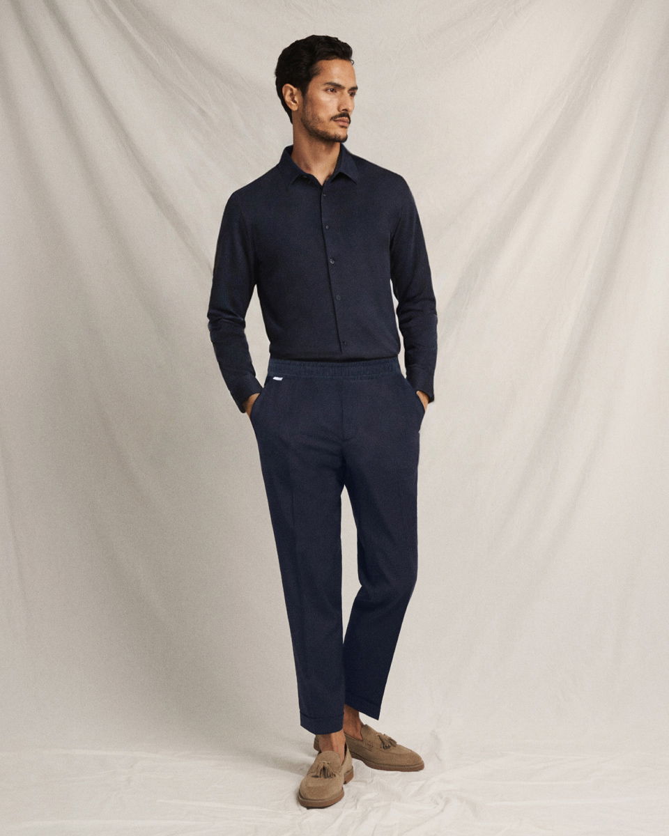 Mann steht vor hellem Hintergrund, trägt dunkelblaue Leinenhose, dunkelblaues Hemd und hellbraune Loafer mit Quasten.