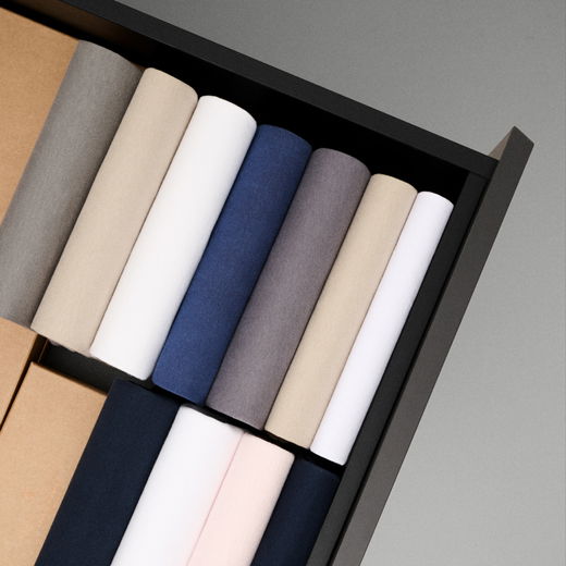 Offene Schublade mit ordentlich eingerollten T-Shirts in verschiedenen Farben wie Beige, Weiß, Blau und Grau, neben Kartons.