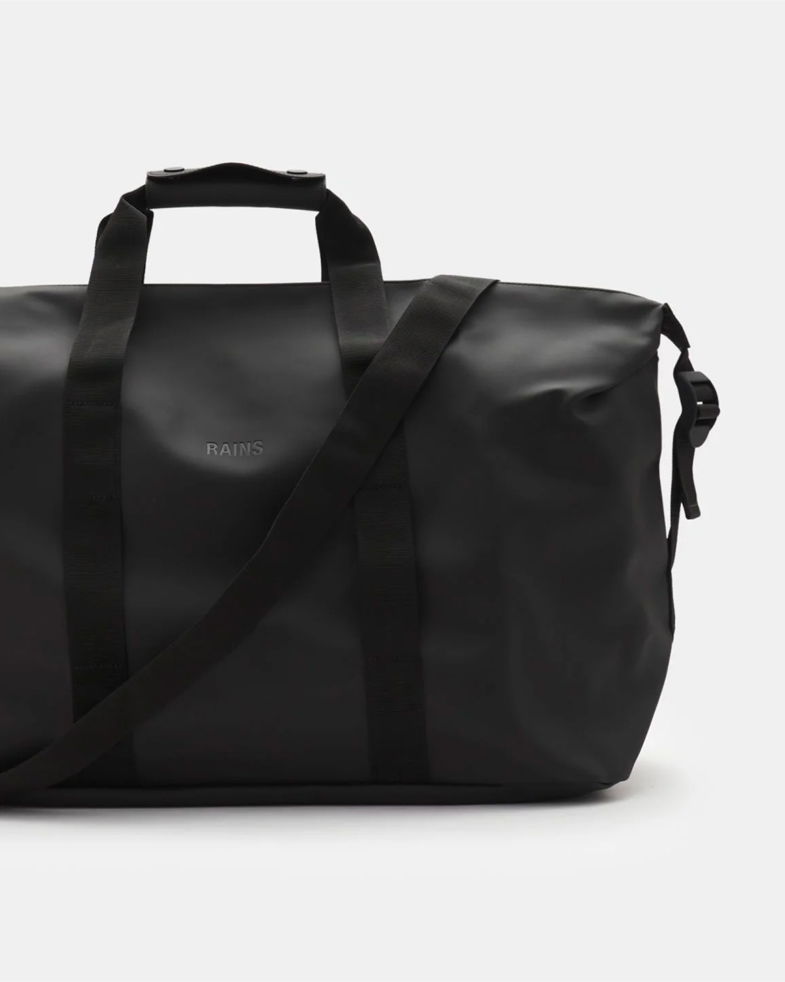 Schwarze Reisetasche aus wasserabweisendem Material mit zwei Tragegriffen, verstellbarem Schulterriemen und RAINS Logo vorne. Schwarze Reisetasche aus wasserabweisendem Material mit zwei Tragegriffen, verstellbarem Schulterriemen und RAINS Logo vorne.