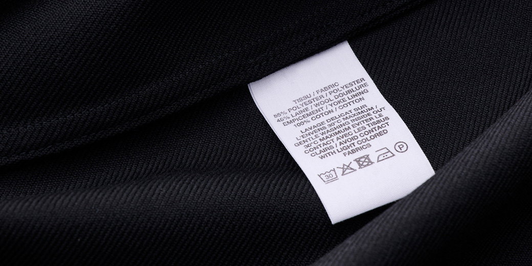 Weißes Waschlabel mit Pflegehinweisen auf dunkelblauem Polyesterstoff, Text und Symbole zur Textilpflege sichtbar.