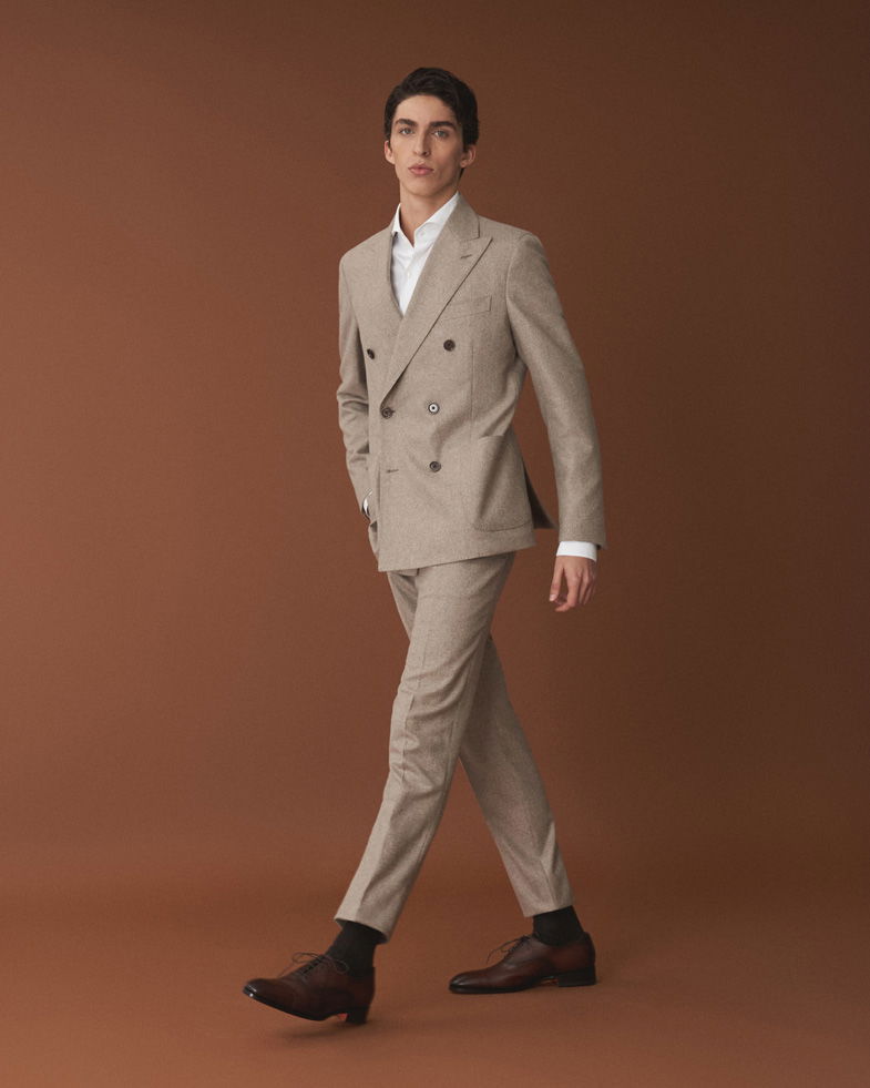 Junger Mann im beigen Anzug mit weißem Hemd, braunen Schnürschuhen und schwarzen Socken vor braunem Hintergrund.