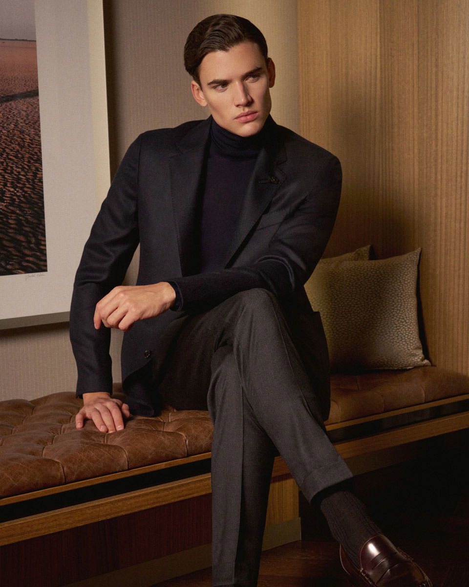 Mann im dunkelblauen Anzug und Rollkragenpullover sitzt auf brauner Ledercouch, trägt braune Loafer, Holzpaneele im Hintergrund. Mann im dunkelblauen Anzug und Rollkragenpullover sitzt auf brauner Ledercouch, trägt braune Loafer, Holzpaneele im Hintergrund.