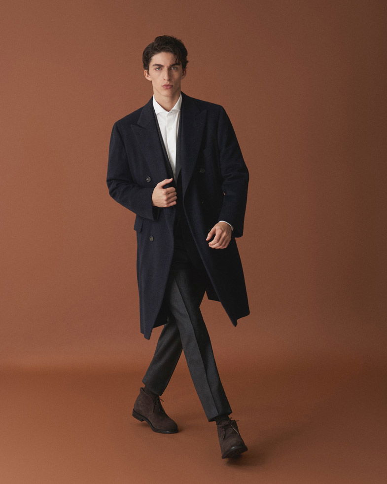 Mann in dunkelblauem Mantel, weißem Hemd, grauer Hose und braunen Schnürstiefeln vor braunem Hintergrund.