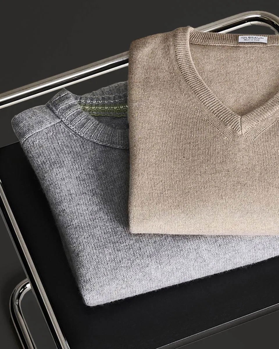 Beige V-Ausschnitt Pullover und grauer Rundhals-Pullover liegen gefaltet auf schwarzem Leder-Hocker mit Metallrahmen.