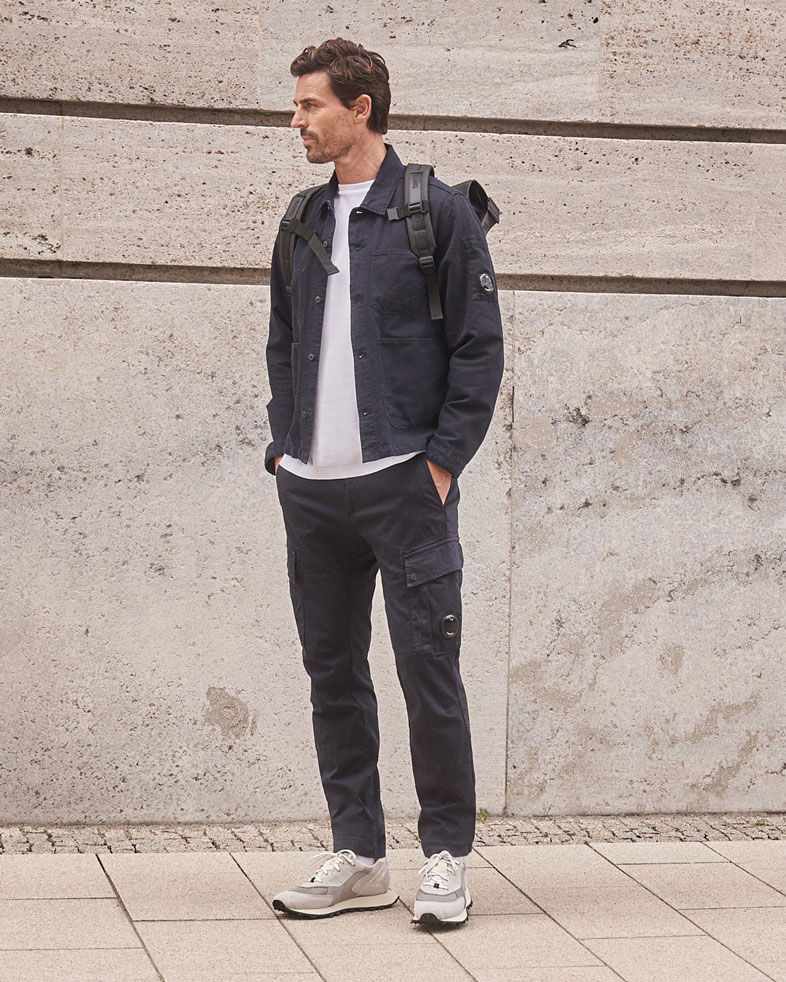 Mann in dunkelblauer Jacke und Cargohose, weißem T-Shirt, grauen Sneakern und schwarzem Rucksack vor Betonwand stehend. Mann in dunkelblauer Jacke und Cargohose, weißem T-Shirt, grauen Sneakern und schwarzem Rucksack vor Betonwand stehend.