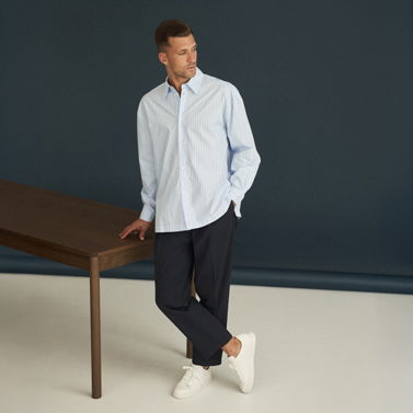 Mann lehnt lässig an Holztisch, trägt hellblau gestreiftes Oversized-Hemd, dunkelblaue Hose und weiße Sneaker. Mann lehnt lässig an Holztisch, trägt hellblau gestreiftes Oversized-Hemd, dunkelblaue Hose und weiße Sneaker.