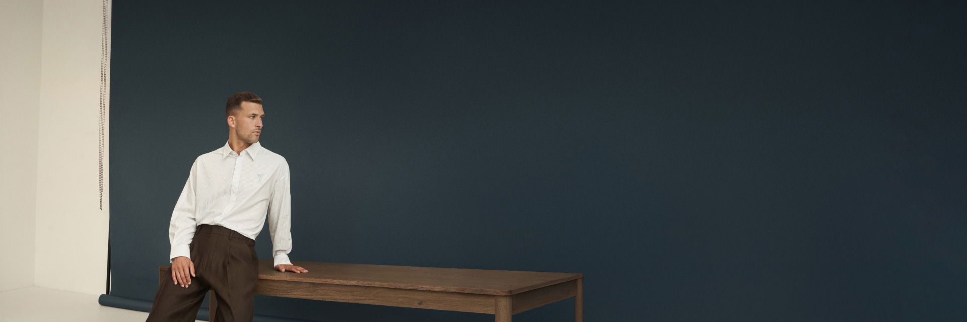 Mann lehnt an braunem Holztisch, trägt weißes Oversized-Hemd und dunkelbraune Hose, vor dunkelblauem Hintergrund. Mann lehnt an braunem Holztisch, trägt weißes Oversized-Hemd und dunkelbraune Hose, vor dunkelblauem Hintergrund.