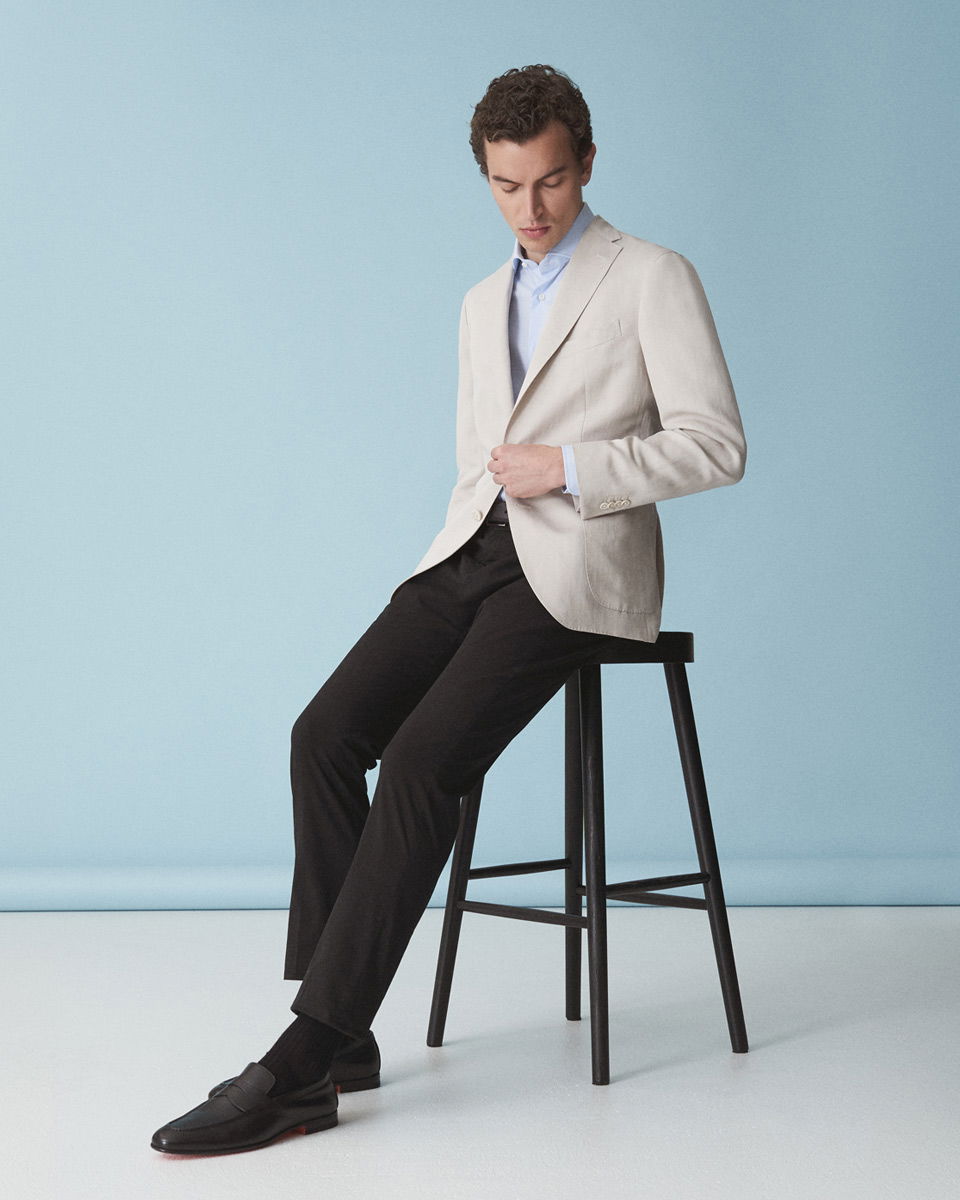 Mann sitzt auf schwarzem Hocker, trägt beiges Sakko, hellblaues Hemd, schwarze Hose und schwarze Loafer, blauer Hintergrund.