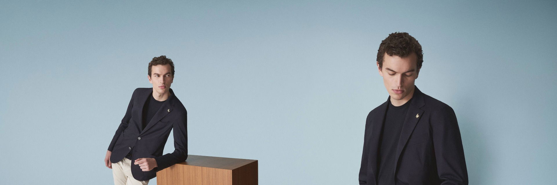 Mann in navy Sakko, schwarzem Shirt und beiger Hose lehnt an Holzsäule, zweite Perspektive zeigt ihn nach unten schauend. Mann in navy Sakko, schwarzem Shirt und beiger Hose lehnt an Holzsäule, zweite Perspektive zeigt ihn nach unten schauend.