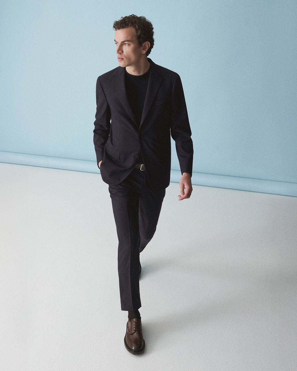 Mann in dunkelblauem Anzug mit schwarzem Rundhalsshirt und braunen Lederschnürschuhen vor hellblauem Hintergrund.