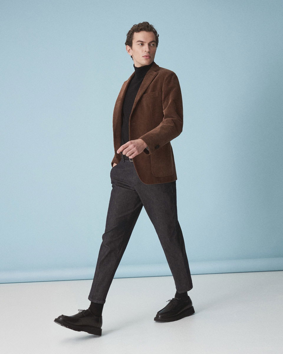 Mann im braunen Cordsakko, schwarzem Rollkragenpullover, grauer Hose und schwarzen Schnürschuhen vor hellblauem Hintergrund.