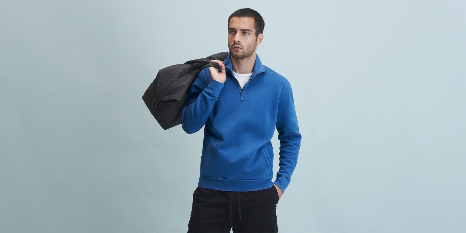 Mann trägt blauen Troyer über weißem T-Shirt, schwarze Hose, hält schwarzen Weekender lässig über der Schulter.