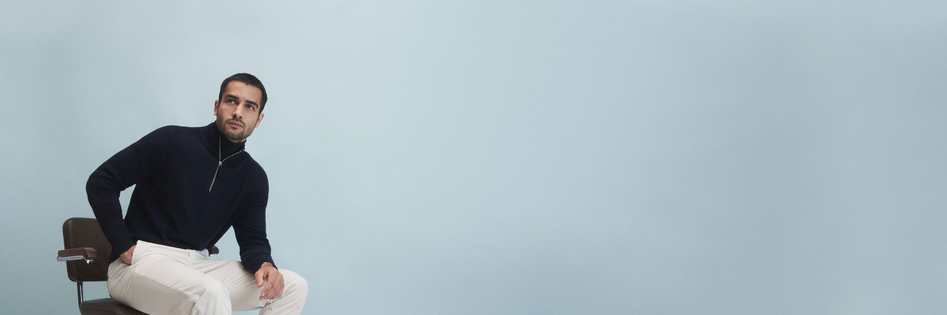 Mann sitzt auf Stuhl, trägt dunkelblauen Troyer mit Reißverschluss über schwarzem Rollkragenpullover und beige Hose vor hellblauem Hintergrund. Mann sitzt auf Stuhl, trägt dunkelblauen Troyer mit Reißverschluss über schwarzem Rollkragenpullover und beige Hose vor hellblauem Hintergrund.