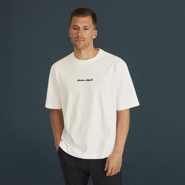 Mann mit kurzem Haar trägt weißes T-Shirt mit schwarzem Schriftzug und dunkelblaue Hose vor dunklem Hintergrund. Mann mit kurzem Haar trägt weißes T-Shirt mit schwarzem Schriftzug und dunkelblaue Hose vor dunklem Hintergrund.