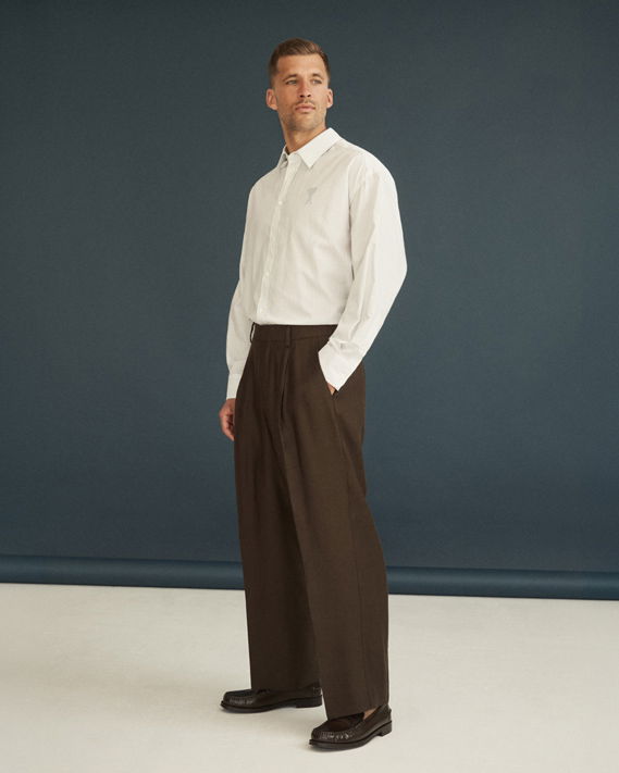 Mann trägt weißen Oversized-Hemd, dunkelbraune weite Hose und braune Loafer, steht vor dunklem Hintergrund. Mann trägt weißen Oversized-Hemd, dunkelbraune weite Hose und braune Loafer, steht vor dunklem Hintergrund.